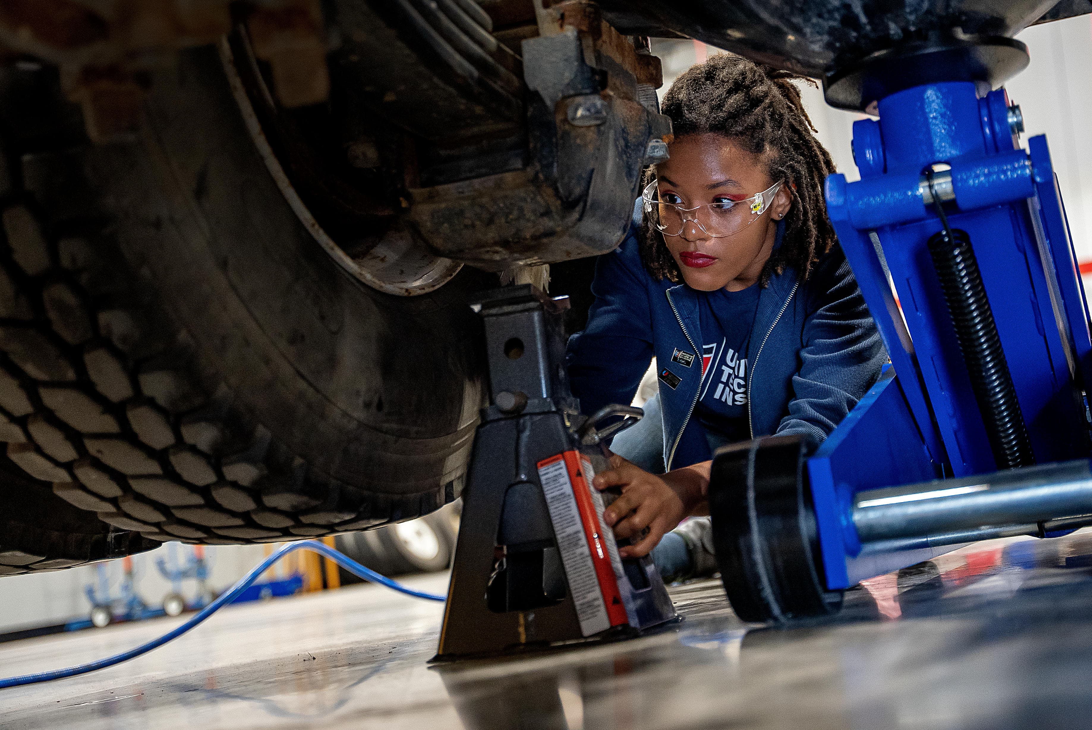 Diesel technician student positioning a jack stand while inspecting a heavy-duty truck.