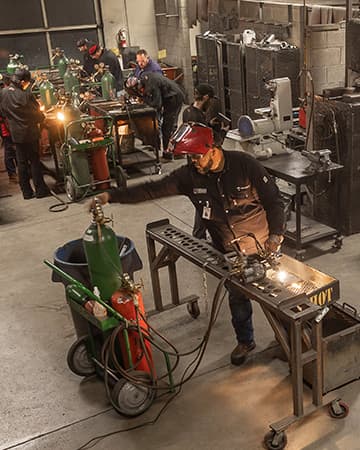 Welding student in protective gear uses a torch, creating sparks while fusing metal pieces.